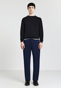 Junger Mann steht aufrecht und trägt einen schwarzen Pullover, marineblaue Hose mit Kordelzug und schwarze Slipper vor einem einfarbigen hellen Hintergrund.