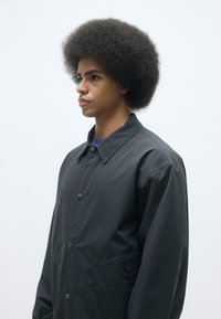 Svart lättviktsjacka med knappstängning fram, spridd krage och sidofickor, bärs över en blå skjorta. Modellen har en voluminös afrofrisyr.