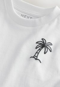 Valkoinen lyhythihainen T-paita, jossa vasemmassa rinnassa musta kirjailtu palmupuu. Resorikaulus ja pehmeä puuvillatekstuuri.