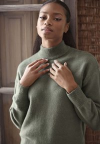 Maglione verde in lana con colletto a imbuto e polsini a coste. Il tessuto appare morbido e testurizzato, aderente in vita. Le mani toccano il colletto.
