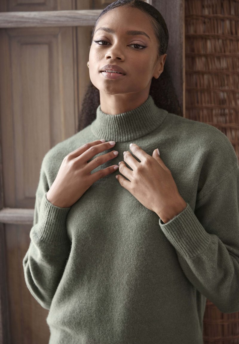 Maglione verde in lana con colletto a imbuto e polsini a coste. Il tessuto appare morbido e testurizzato, aderente in vita. Le mani toccano il colletto.