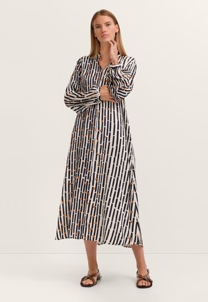 Woman standing with hand on chin wearing a long-sleeve, button-up midi dress with black vertical stripes and small orange dots, and brown sandals.