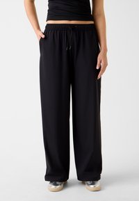 Pantalon large noir avec taille élastique et cordon de serrage, doté de poches latérales. Tissu lisse, léger avec une coupe décontractée.