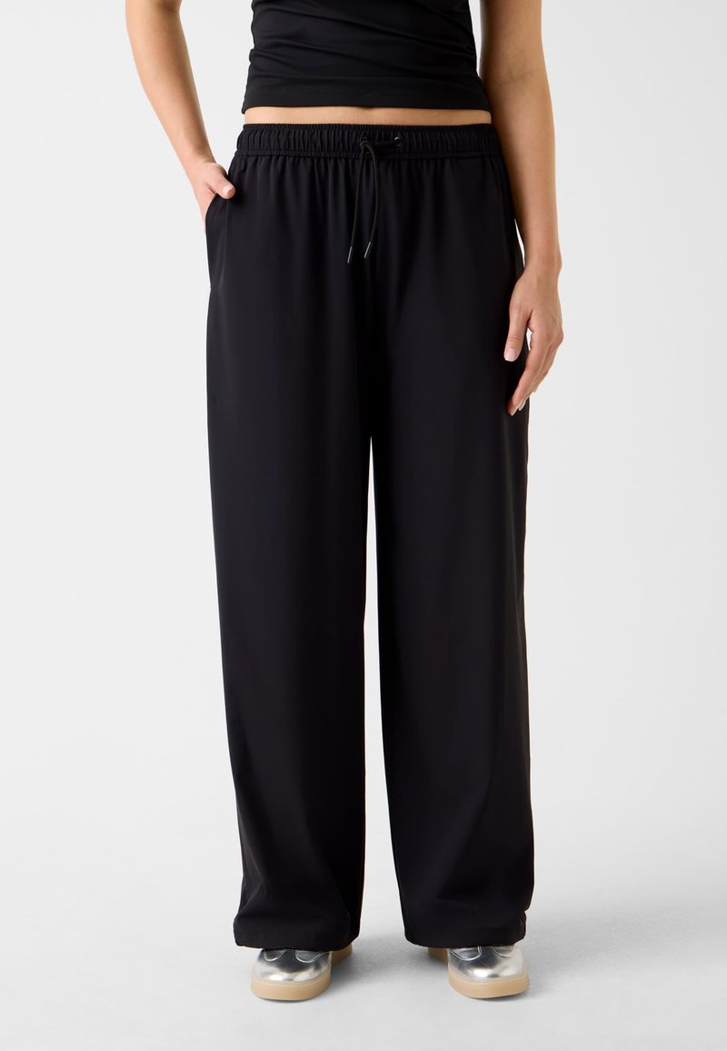 Pantalon large noir avec taille élastique et cordon de serrage, doté de poches latérales. Tissu lisse, léger avec une coupe décontractée.