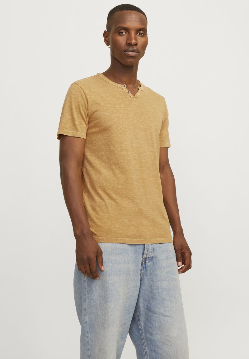 Camisa de manga corta de color miel con cuello en V y dos botones cerca del cuello. La tela parece suave con una ligera textura.