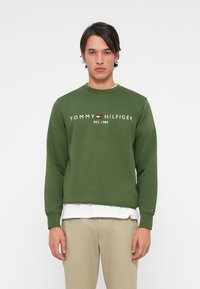 Green cotton sweatshirt featuring a crew neck, ribbed cuffs, and the text "TOMMY HILFIGER EST. 1985" in white. Logo accents on sleeves.