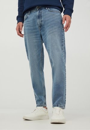 Person trägt hellblaue Jeans mit geradem Schnitt, weiße Turnschuhe und ein dunkelblaues Langarmshirt, steht vor einem einfarbigen Hintergrund.