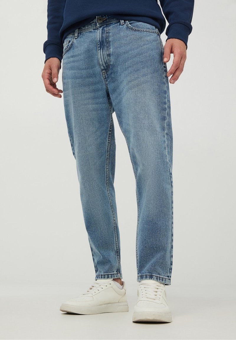 Personne portant un jean droit bleu clair, des baskets blanches et un haut à manches longues bleu marine, debout devant un fond uni.