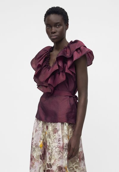 Γυναίκα που φοράει μπορντό τοπ χωρίς μανίκια με μεγάλα βολάν και ημιδιάφανο φούστα με λουλουδένιο μοτίβο σε απλό γκρι φόντο.