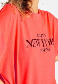 Nainen, joka pukeutuu kirkkaan korallinväriseen ylisuureen t-paitaan, jossa on mustalla etupuolella teksti "New York" ja maantieteelliset koordinaatit.