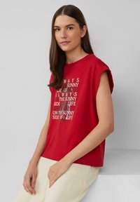 Rode t-shirt met korte mouwen en een ronde hals, met daarop een witte en zwarte tekstafdruk in een gelaagd ontwerp aan de voorkant, gecombineerd met lichtbeige broek.