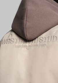 Hættejakke i lys beige med et struktureret, glat stof. Har en kontrastfarvet brun hætte og prægede logo-detaljer på ryggen.