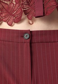 Pantalones burdeos con rayas finas, con un diseño de frente plano, cierre con botón negro y tela texturizada que muestra una blusa de encaje floral encima.