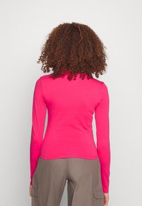 Osoba s kudrnatými hnědými vlasy, oblečená v jasně růžovém tričku s dlouhým rukávem a šedých cargo kalhotách, stojí zády k objektivu na neutrálním pozadí.