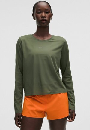 Mujer con una camiseta de manga larga verde oliva y pantalones cortos deportivos naranja brillante sobre un fondo claro y liso.