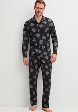 Mann trägt passende dunkle Pyjamas mit Sonnen- und Wellenmustern, steht barfuß vor einer schlichten hellen Wand.