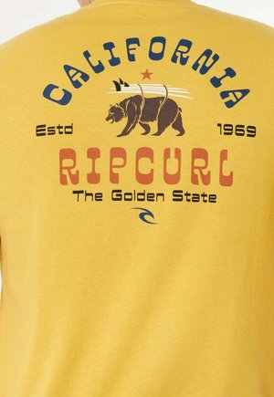 T-shirt en coton jaune avec un imprimé graphique représentant un ours, des planches de surf, ainsi que les textes "California" et "Ripcurl" en couleurs contrastées.