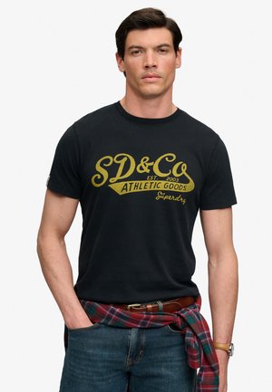 Mand iført sort t-shirt med gul tekst ""SD&Co Athletic Goods"", blå jeans, brun bælte, rød ternet skjorte bundet om livet og et armbåndsur.