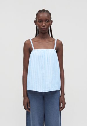 Γυναίκα με πλεξούδες, που φοράει ένα αμάνικο τοπ με ανοιχτό μπλε ριγέ μοτίβο και φαρδιά σκούρα μπλε παντελόνια, στέκεται μπροστά σε απλό φόντο.