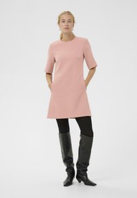 Rosa kurzärmeliges Kleid mit geradem Schnitt und Seitentaschen, getragen mit schwarzen kniehohen Stiefeln und schwarzen Strumpfhosen vor einem schlichten Hintergrund.