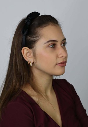 Czarna materiałowa opaska na głowę z węzłowym detalem. Noszona z złotymi kolczykami kołami. Osoba ma długie, proste włosy i nosi bordowy top.