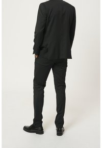 Completo nero su misura con giacca strutturata e spacchetto centrale, abbinato a pantaloni slim-fit e scarpe nere lucide.