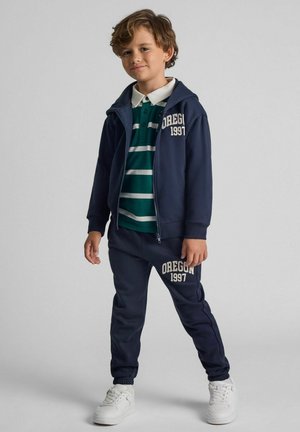 Navy tracksuit set featuring zip-up hoodie and elasticated pants with "OREGON 1997" print. Striped green polo shirt underneath. White sneakers.