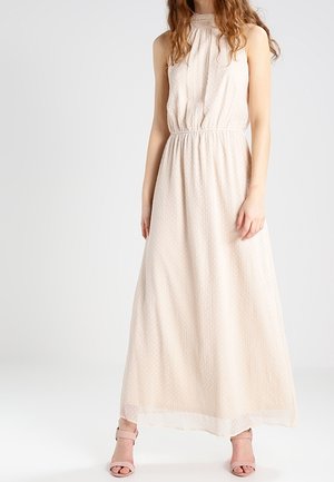 Kobieta w bezrękawowej, o wysokim dekolcie, jasnobeżowej sukience maxi z subtelnym wzorem kropek oraz pasujących sandałach na obcasie, stojąca na jednolitym tle.