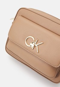 Bežová kožená kabelka na rameno Calvin Klein s přední kapsou a zlatým detailem loga CK na bílém pozadí.