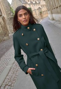 Tamnozelena dvoredna jakna s zlatnim gumbima, visokim ovratnikom i remenom, izrađena od glatkog materijala; ima bočne džepove i prilagođen kroj.