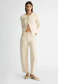 Giacca cropped beige chiaro con bordi sfilacciati, abbinata a pantaloni larghi coordinati. Il modello indossa ballerine dorate con punta affilata.