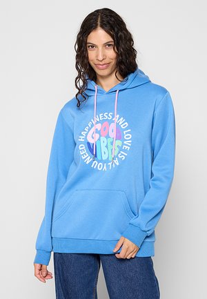 Woman with long curly hair wears a blue hoodie with “Good Vibes” and positive message on front, standing and smiling at the camera.