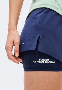 Main ajustant un short de course bleu marine sous une jupe fendue bleue avec le texte « LONDON 42.195KM 26.219MI » sur le short.
