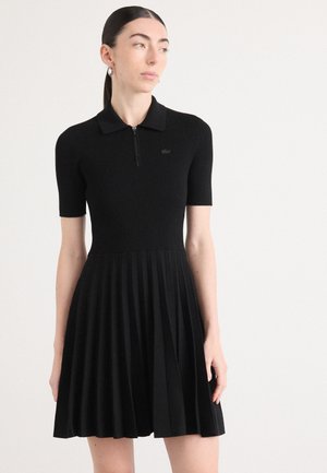 Black ribbed dress with a polo collar, short sleeves, a front zipper, and pleated skirt. Features a small, embossed logo on the chest.