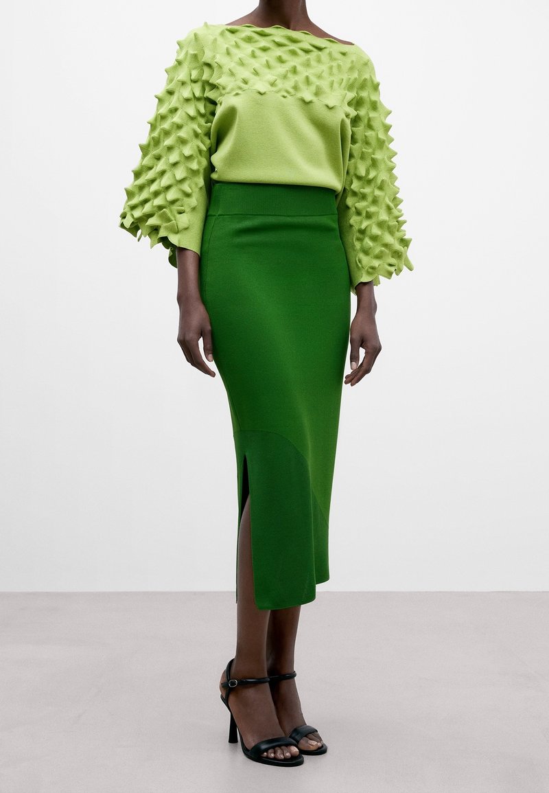 Mannequin portant un pull texturé vert clair avec des motifs en relief et une jupe midi taille haute vert foncé avec une fente à l'avant.