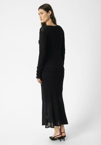 Abito nero in maglia con maniche lunghe, caratterizzato da dettagli testurizzati e orlo svasato. Indossato con tacchi neri alti. Finitura liscia e elegante.