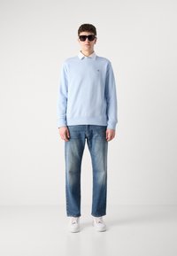 Homme portant un sweat-shirt bleu clair par-dessus une chemise blanche à col, un jean bleu, des baskets blanches et des lunettes de soleil noires, debout devant un fond uni.