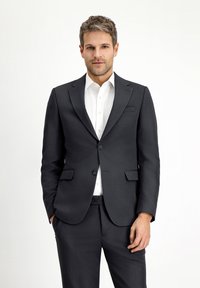 Abito grigio scuro con giacca attillata, revers a punta e due bottoni; abbinato a una camicia bianca, tessuto liscio e minimalismo nei dettagli.