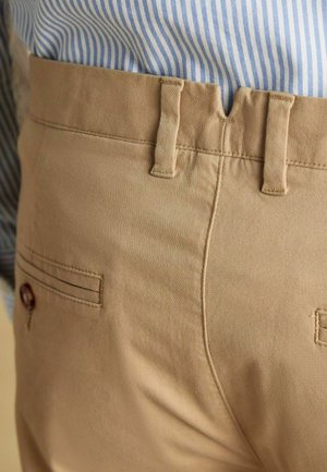 Beige Baumwollhosen mit Detail an der Gesäßtasche, sichtbaren Nähten und Gürtelschlaufen. Gestreiftes blau-weißes Hemd teilweise sichtbar darüber.