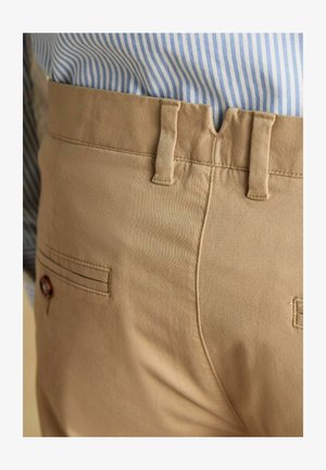 Beige Baumwollhosen mit Detail an der Gesäßtasche, sichtbaren Nähten und Gürtelschlaufen. Gestreiftes blau-weißes Hemd teilweise sichtbar darüber.