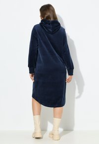 Navy hupparimekko, joka on valmistettu pehmeästä, plush-materiaalista. Mekossa on pitkät hihat ja rento malli, joka ulottuu keskilevylle, ja se on yhdistetty beigeihin saappaisiin.