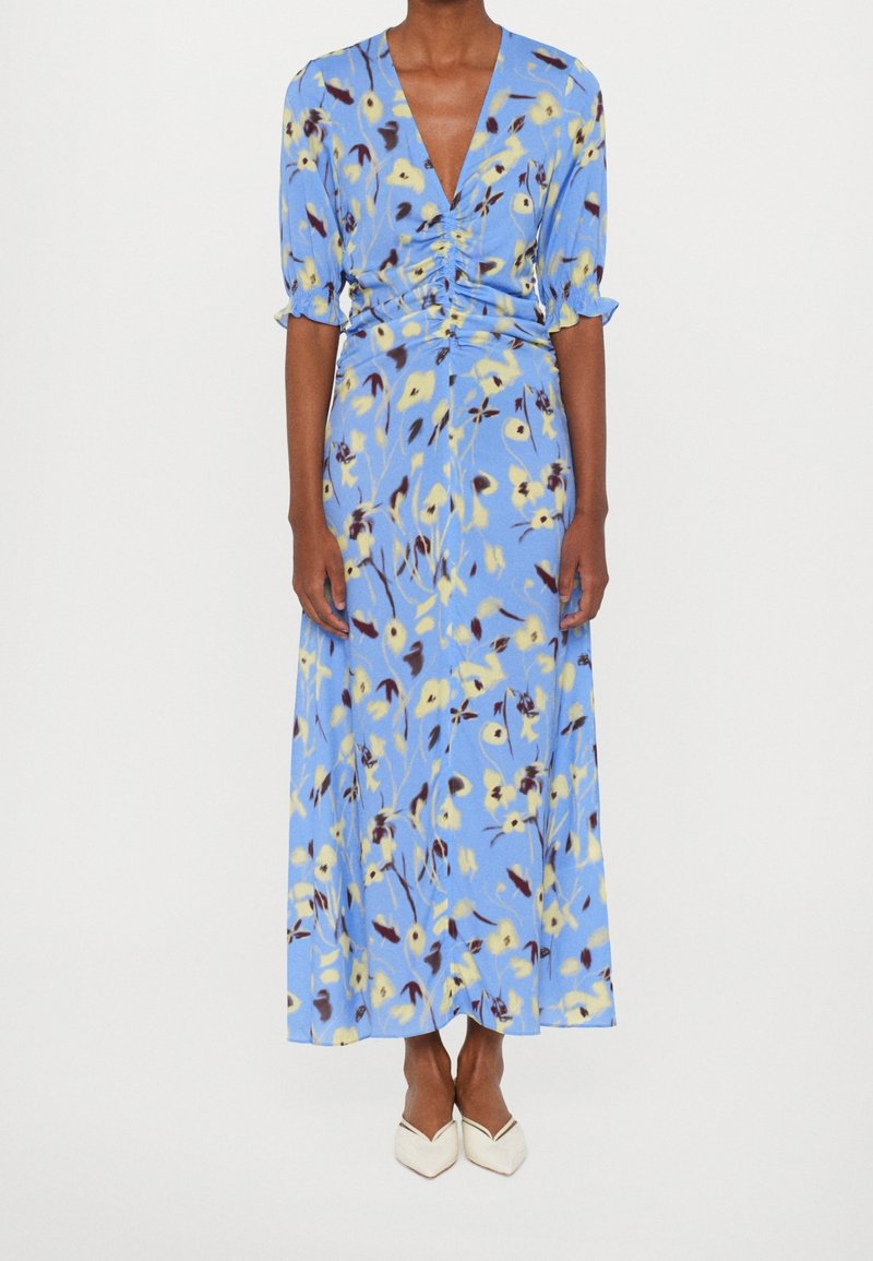 Femme portant une robe midi fleurie bleu clair avec un devant froncé, des manches trois-quarts et des talons blancs pointus fond blanc.