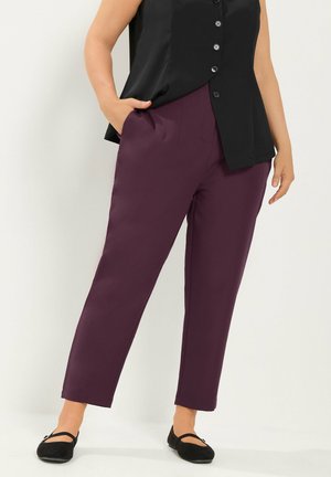 Mujer con pantalones cónicos de color púrpura oscuro, top negro sin mangas con botones y zapatos planos Mary Jane negros, de pie con una mano en el bolsillo.