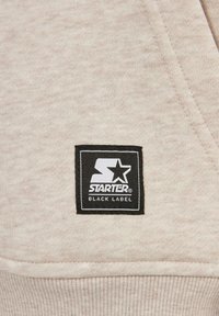 Beige sweatshirtstoff med teksturert overflate. Har et svart firkantet merke med hvit "STARTER"-logo og "BLACK LABEL"-tekst.