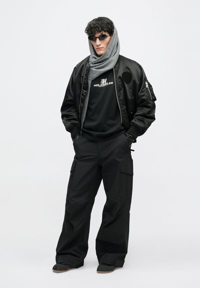 Vyras, kuris dėvi juodus akinius nuo saulės, juodą bombonešio tipo striukę, juodas krovinių kelnes, juodas pirštines ir pilką šaliką, apvyniotą aplink galvą.