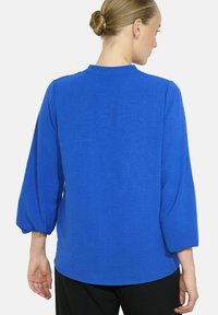 Woman with blonde hair in a bun wearing a bright blue long-sleeve blouse and black pants, shown from the back against a white background.