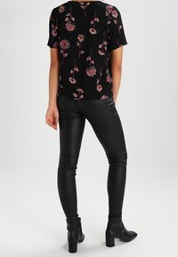 Woman wearing black floral short-sleeve top, black fitted leather pants, and black heeled ankle boots, standing facing away.