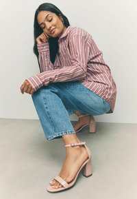Sandali con tacco in pelle lucida rosa, con una fascia ampia e fibbia alla caviglia, abbinati a jeans a gamba dritta azzurri e una camicia a righe.