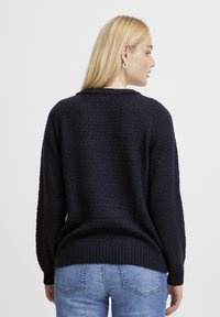 Marineblauer Strickpullover mit einem strukturierten Muster, rundem Ausschnitt sowie elastischem Saum und Ärmeln. Getragen mit hellblauen Jeans.