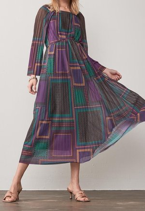 Femme portant une robe longue à manches longues plissée avec de grands motifs carrés géométriques violet, vert et marron, associée à des sandales à talons beiges.